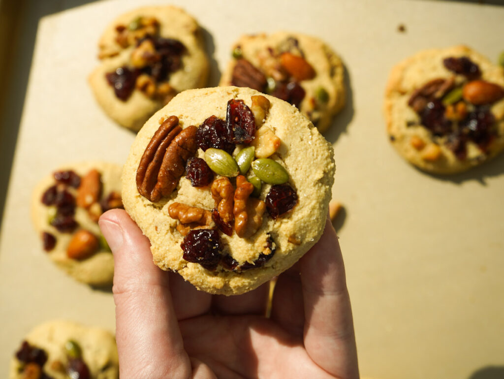A hand holding freshly baked trail mix cookie above a gold baking tray with the other cookies on it.