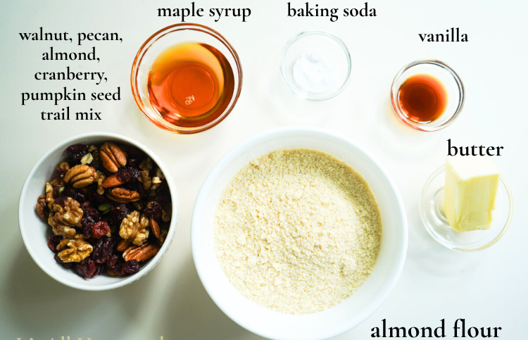 A flat-lay image of baking ingredients for almond flour trail mix cookies: maple syrup, almond flour, vanilla, baking soda, butter, and a cup of trail mix.
