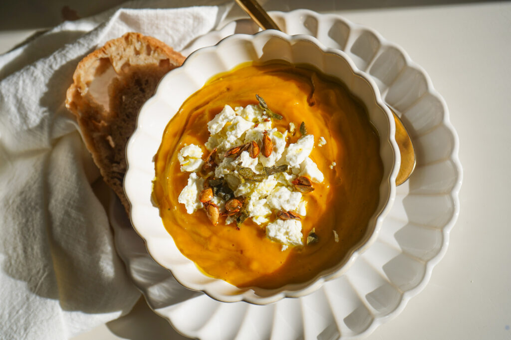 A white scalloped bowl with creamy butternut squash soup topped with crumbled feta, roasted seeds, and crispy sage.