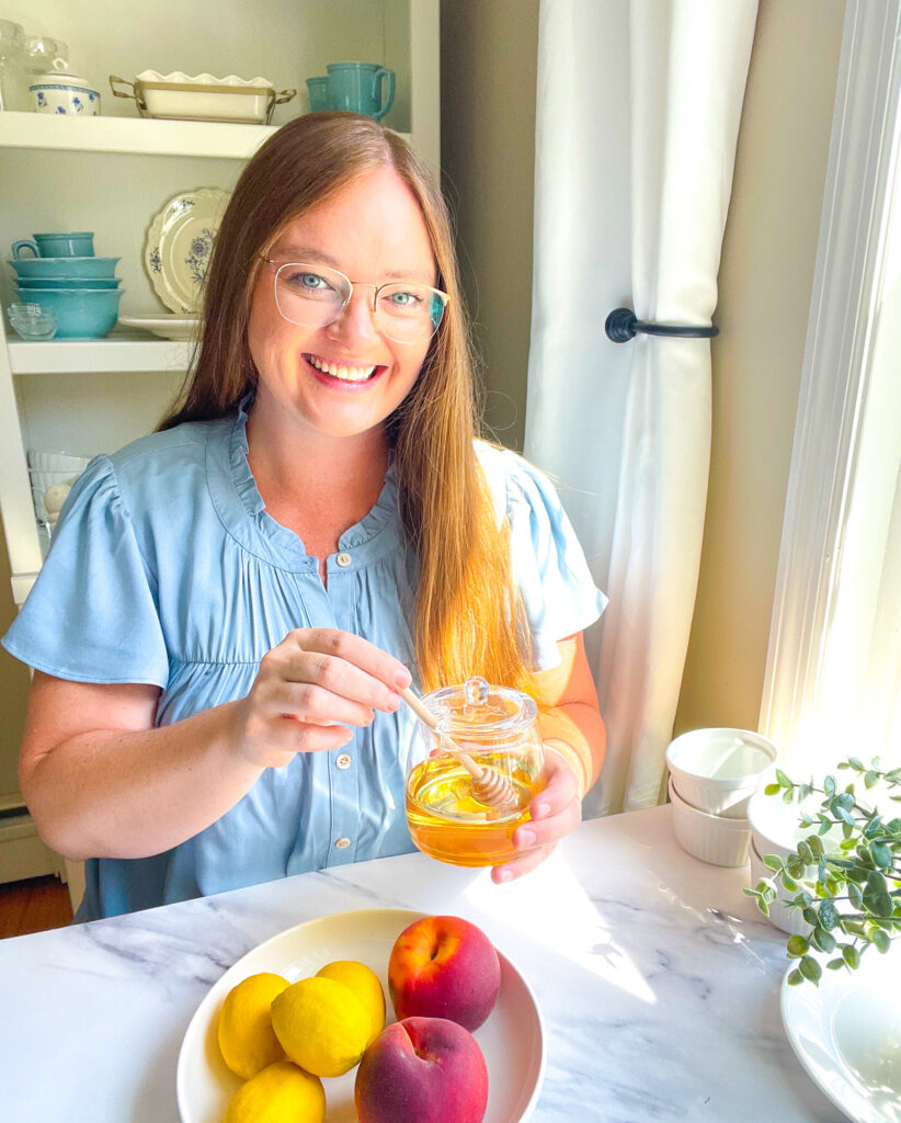 a woman with long brown hair wearing a blue blouse sitting at a marble table with a honey jar and a bowl of lemons and peaches
