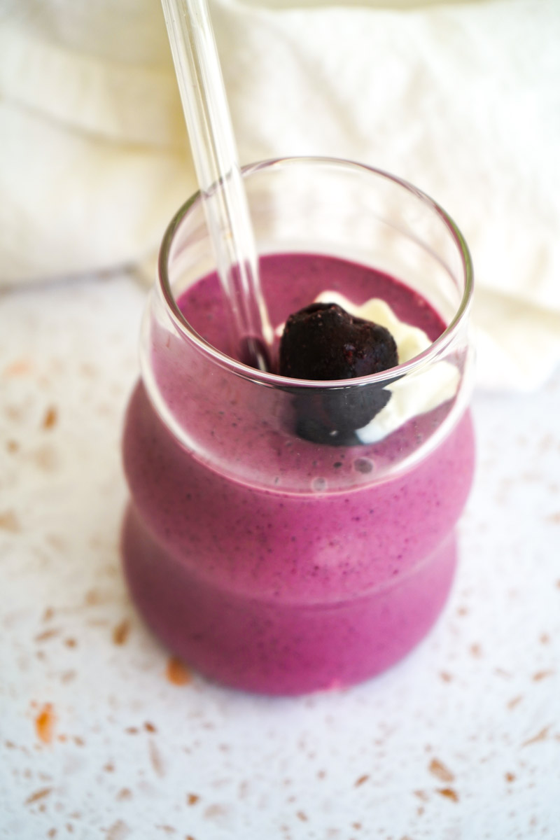 cherry berry smoothie in a curvy glass with a straw topped with a frozen cherry and greek yogurt, on a terrazzo counter