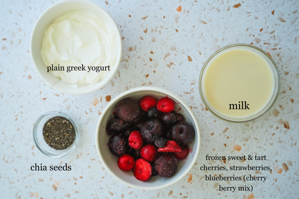 ingredients for a protein cherry berry smoothie: plain greek yogurt, milk, berries, and chia seeds on a white terrazzo counter.