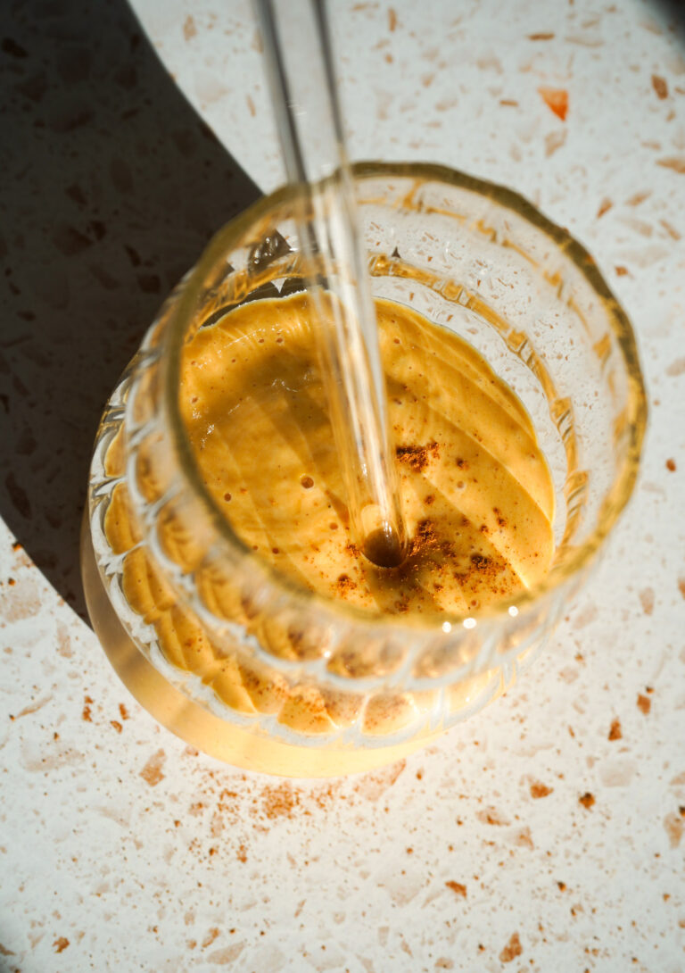 Pumpkin Vanilla Smoothie in a curvy glass with a glass straw and sprinkle of cinnamon on top, on a white terrazzo counter.