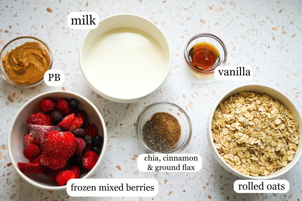 a flat lat of ingredients for PB & J Oatmeal in bowls: frozen mixed berries, rolled oats, vanilla, chia seeds, flax, cinnamon, milk, maple syrup and PB.