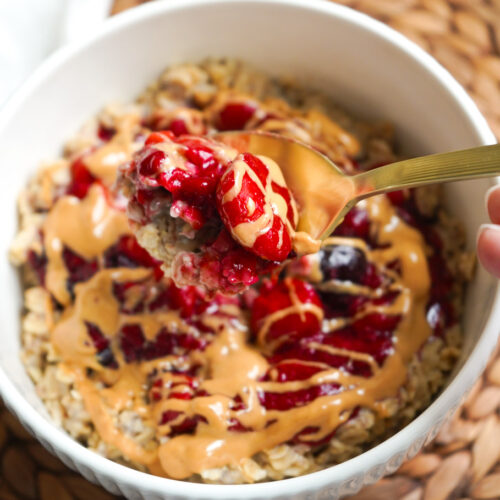 a white fluted bowl with PB & J Oatmeal in it, a gold spoon showing the texture, ready to take a bite.
