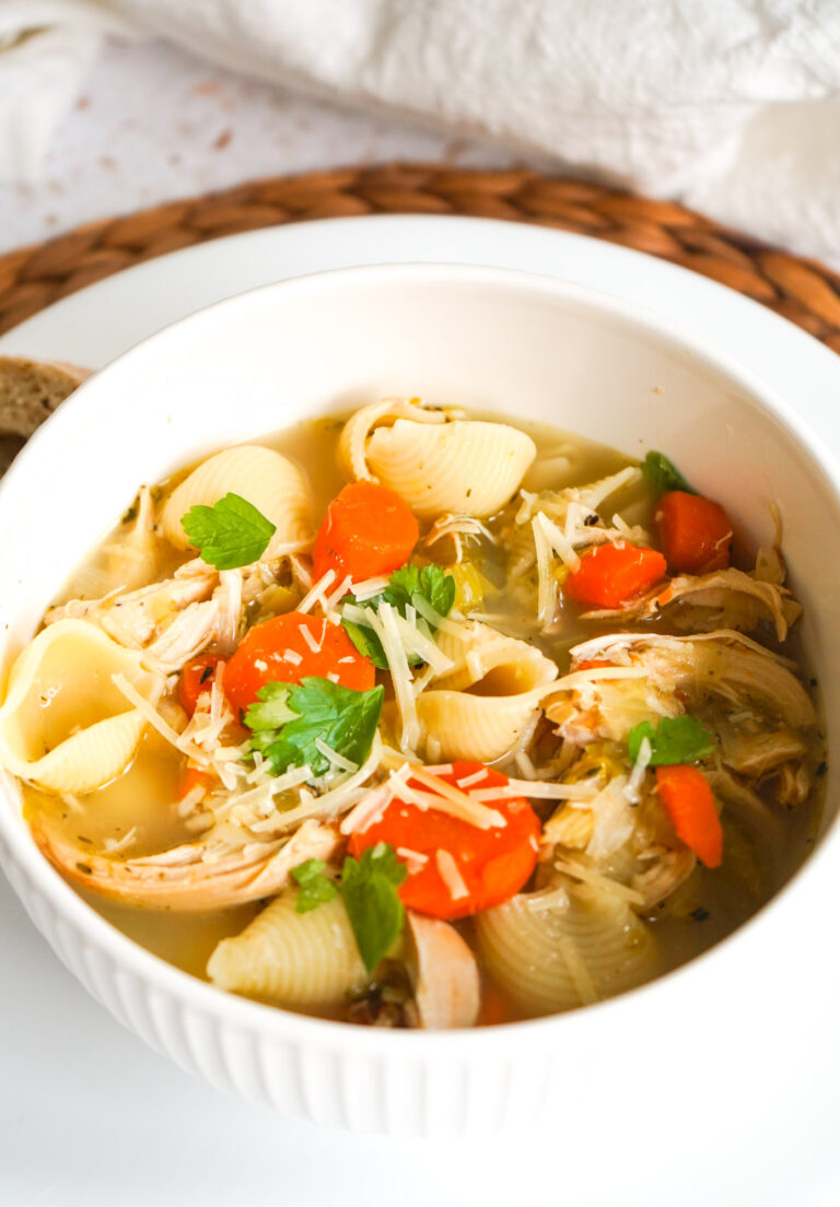 Cozy bowl of chicken noodle soup served in a white fluted bowl, topped with fresh parsley, a sprinkle of parmesan, and a drizzle of olive oil on top, with a linen napkin and bread on the side.