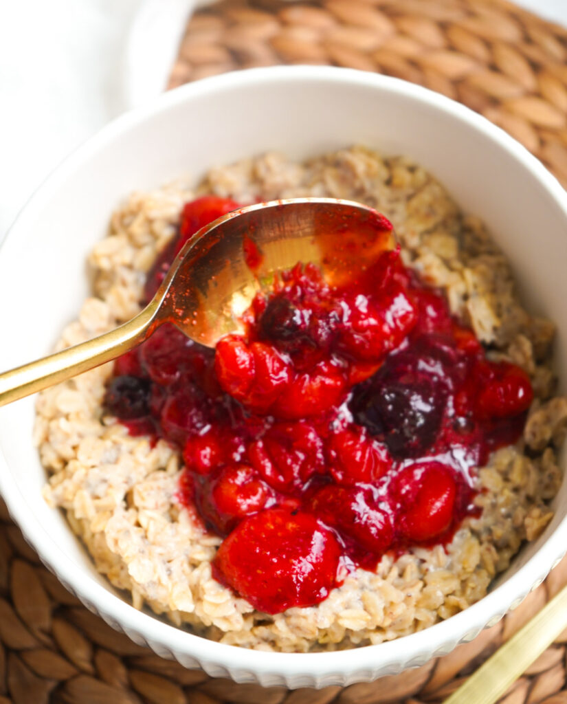 A gold spoon spooning warm berry mixture onto fluffy cooked oatmeal in a white bowl.