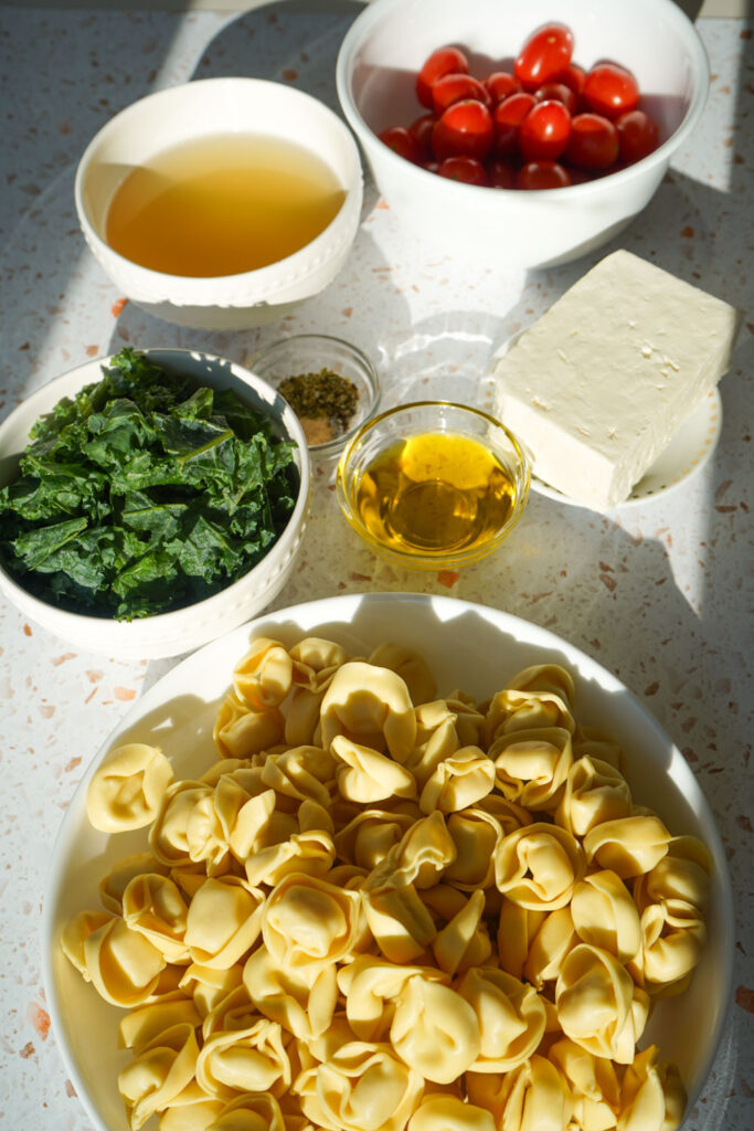 flat lay of ingredients in bowls for chicken tortellini bake: cheese tortellini, spinach, olive oil, seasonings, broth, tomatoes feta.