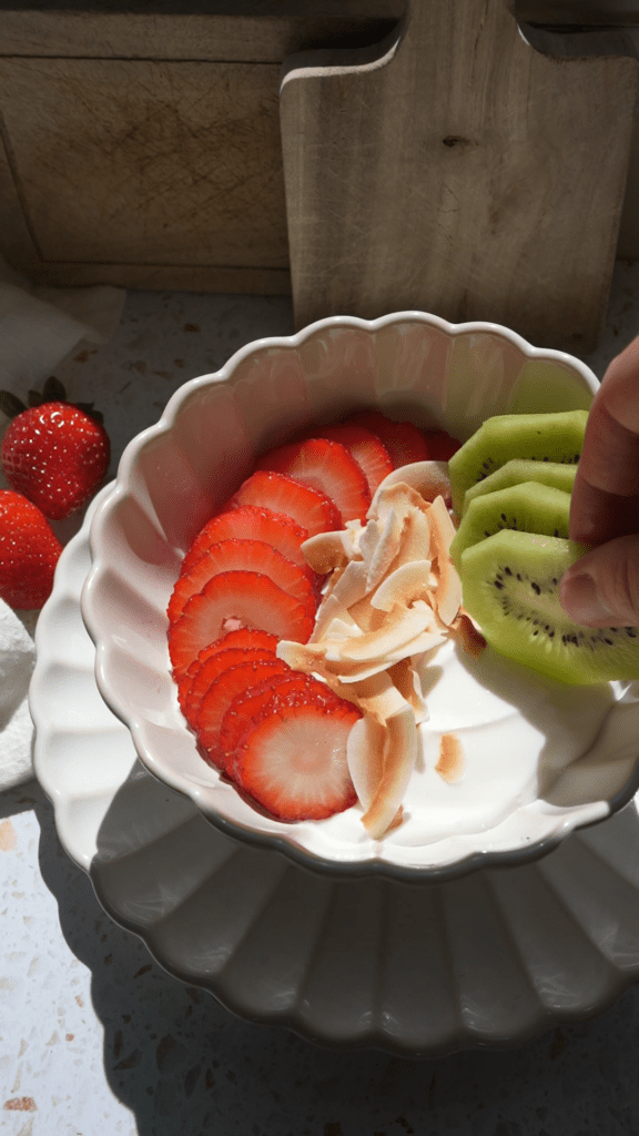 Hand adding kiwi slices to a strawberry kiwi coconut yogurt bowl.