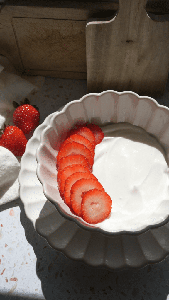 Adding sliced strawberries in a row on vanilla Greek yogurt in a white scalloped bowl.