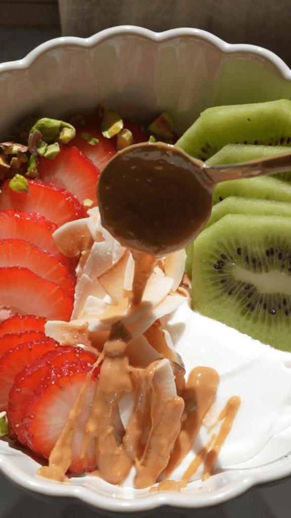 Adding natural peanut butter drizzle to strawberry kiwi yogurt bowl with a small gold spoon.