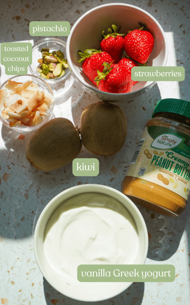 A flat lay of ingredients for a strawberry kiwi yogurt bowl: vanilla greek yogurt, natural peanut butter, kiwi, strawberries, toasted coconut chips, and pistachios.