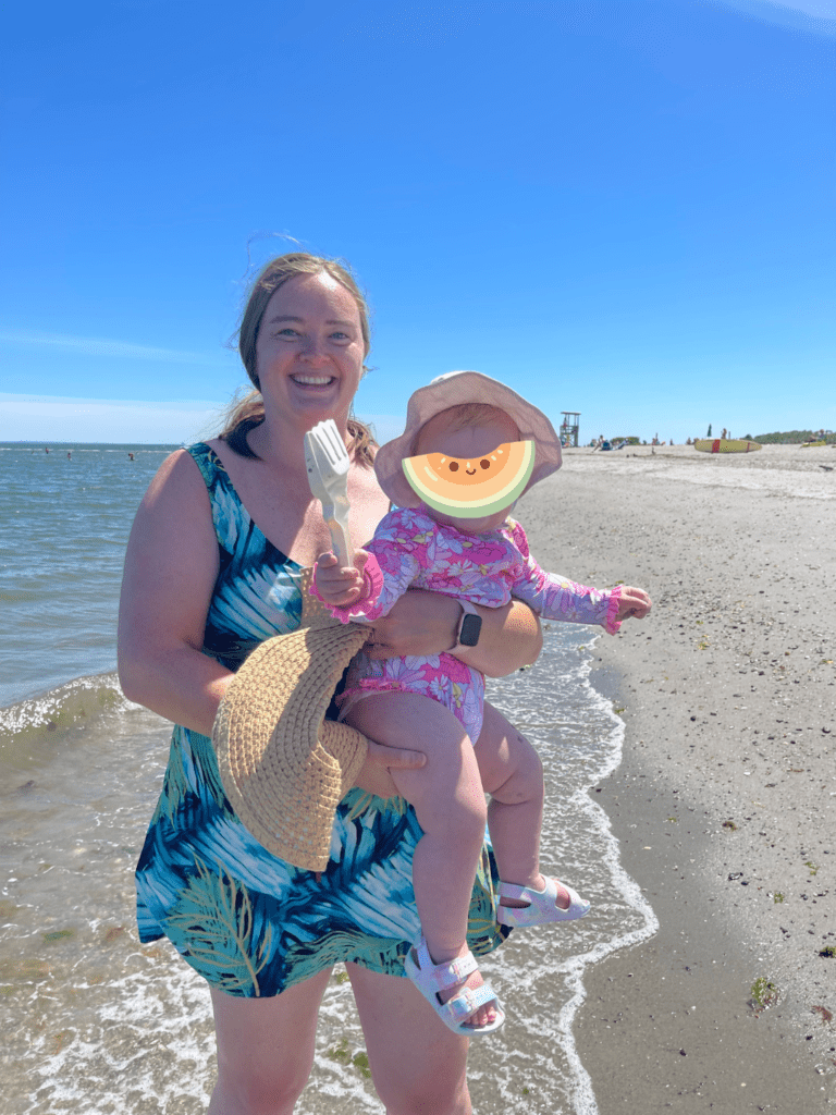 A woman smiling in a blue floral swim dress standing at a shoreline holding a toddler in a pink floral swimsuit and a toy shovel in hand, child's face covered with a cantaloupe emoji.