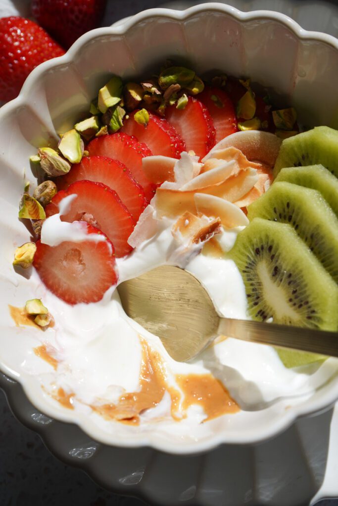 A yogurt bowl with sliced strawberries, kiwi, chopped pistachios and coconut flakes with a drizzle of peanut butter in a white scalloped bowl.