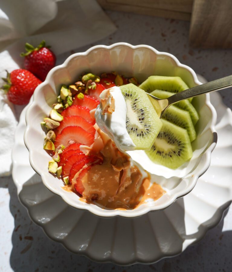A yogurt bowl with sliced strawberries, kiwi, chopped pistachios and coconut flakes with a drizzle of peanut butter in a white scalloped bowl.