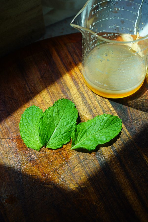 three bright green mint leaves on a cutting board in the sunlight with a glass mixing jar with honey and lime juice to make a fruit salad dressing