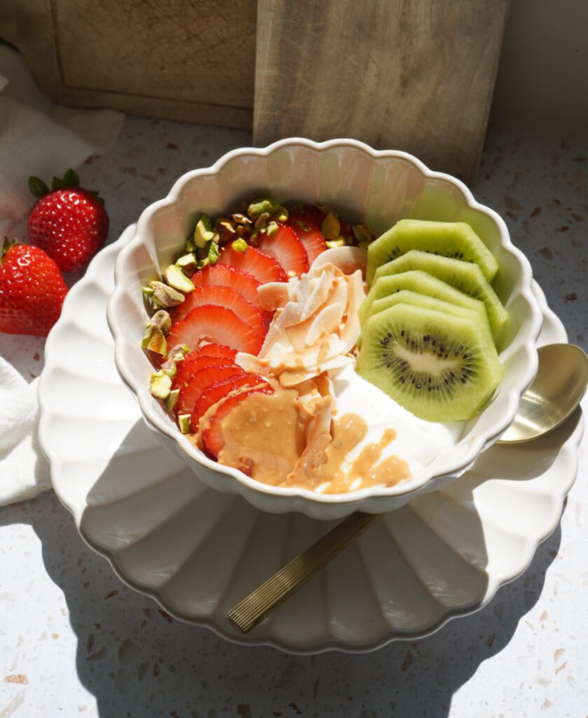 A yogurt bowl with sliced strawberries, kiwi, chopped pistachios and coconut flakes with a drizzle of peanut butter in a white scalloped bowl.