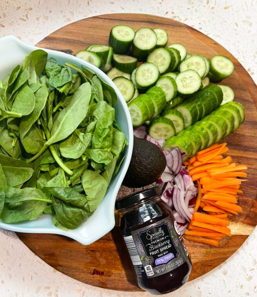 Fresh ingredients for blackberry grilled chicken salad on an acacia wood board including spinach, cucumber, carrots, sliced red onion, avocado and blackberry preserves.