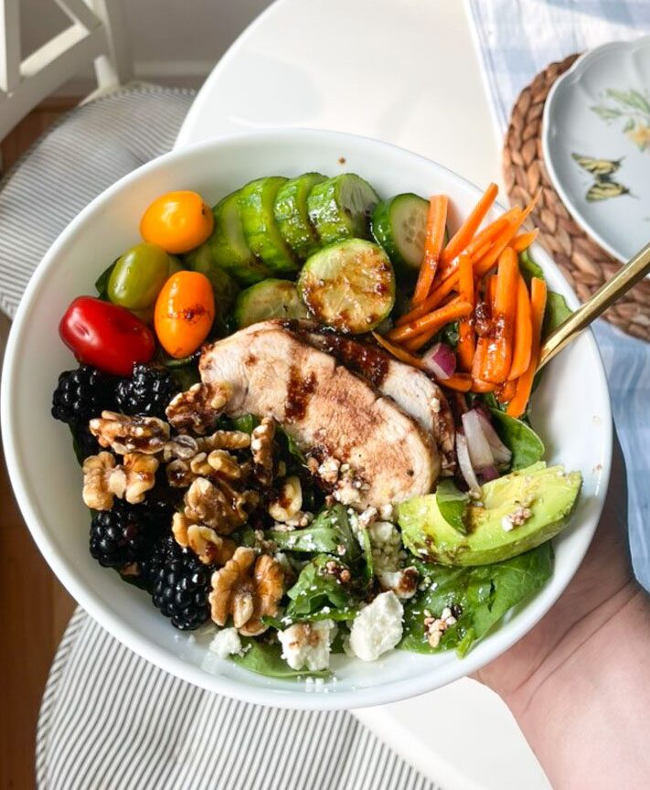 hand holding a white bowl full of a colorful blackberry chicken salad with grilled chicken, carrots, tomatoes, cucumber, walnuts avocado, spinach and feta topped with blackberry balsamic dressing.