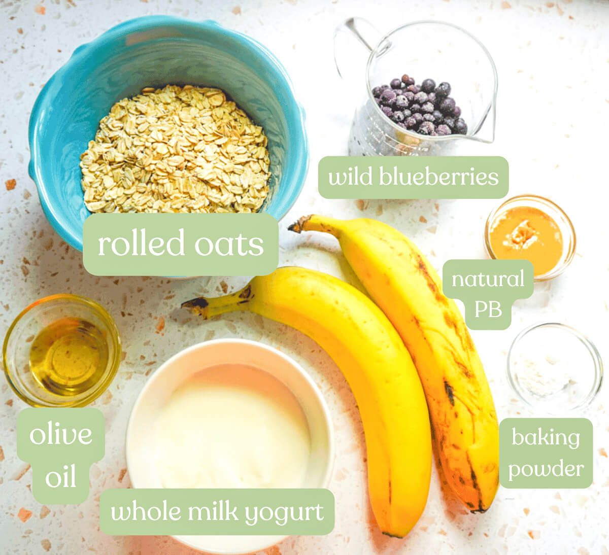 Flat lay ingredients for soft baked wild blueberry banana oat fingers including rolled oats in a teal bowl, two ripe bananas, frozen wild blueberries, whole milk yogurt, olive oil, peanut butter and baking powder on a white terrazzo surface