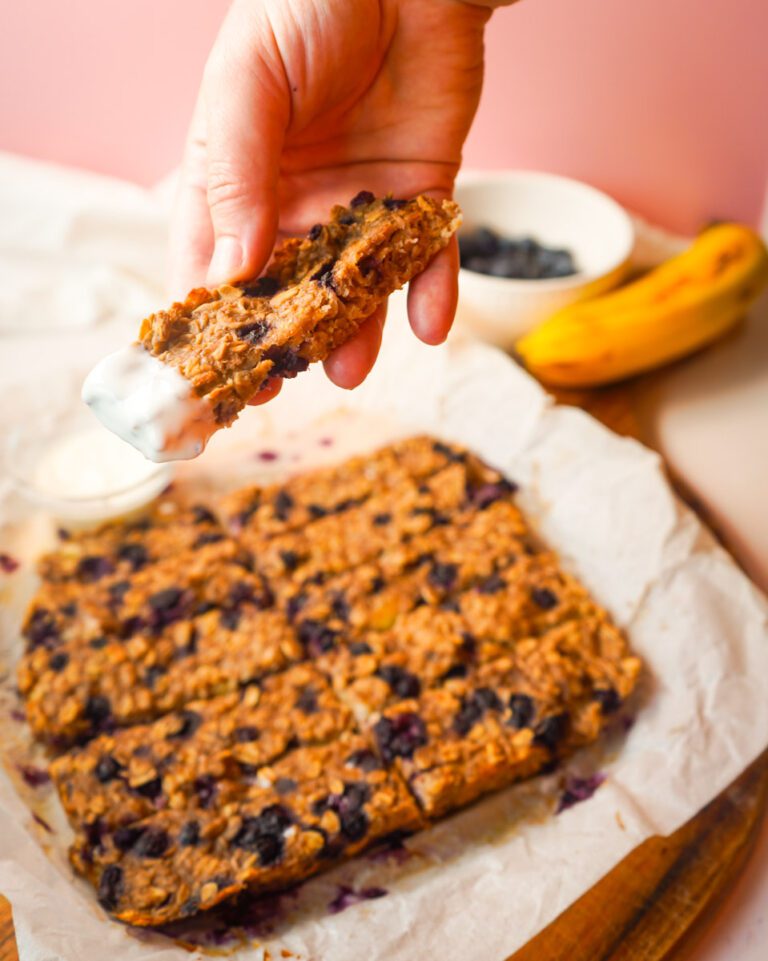 Hand lifting a soft blueberry banana oat finger from parchment paper with a small bowl of Greek yogurt for dipping and banana and blueberries in the background