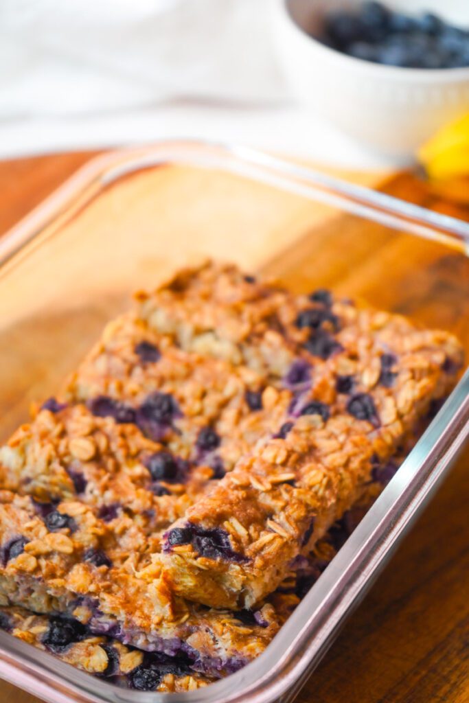 Soft baked blueberry banana oat bars cut into fingers stored in a glass meal prep container on a wooden board with blueberries and banana in the background