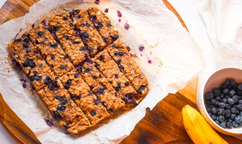 Soft baked wild blueberry banana oat fingers cut into strips on parchment paper on a wooden board with a bowl of blueberries and banana alongside