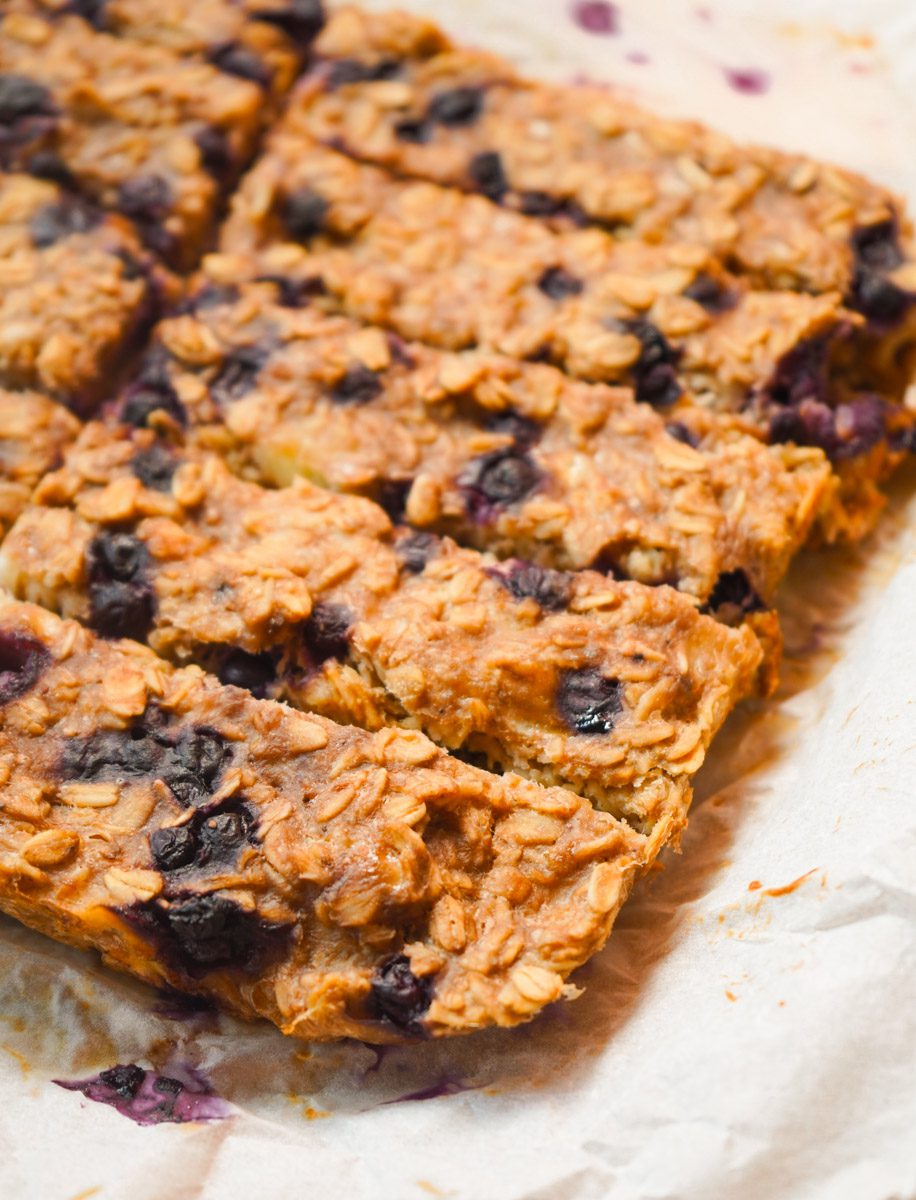 Close up of soft baked blueberry banana oatmeal bars cut into finger strips on white parchment showing golden oat texture and wild blueberries