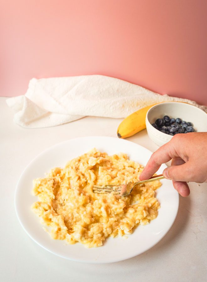 Hand mashing two ripe bananas with a gold fork on a white plate with a bowl of blueberries and banana in the background on a pink backdrop
