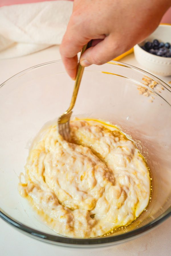 Hand stirring wet ingredients including mashed banana yogurt and olive oil together in a large glass bowl with a gold fork with blueberries in the background