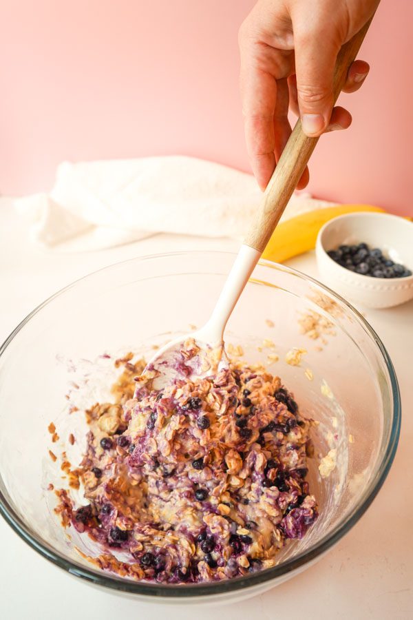 Hand folding frozen wild blueberries and rolled oats into banana yogurt batter in a glass bowl with a wooden spatula with banana and blueberries in background on pink backdrop