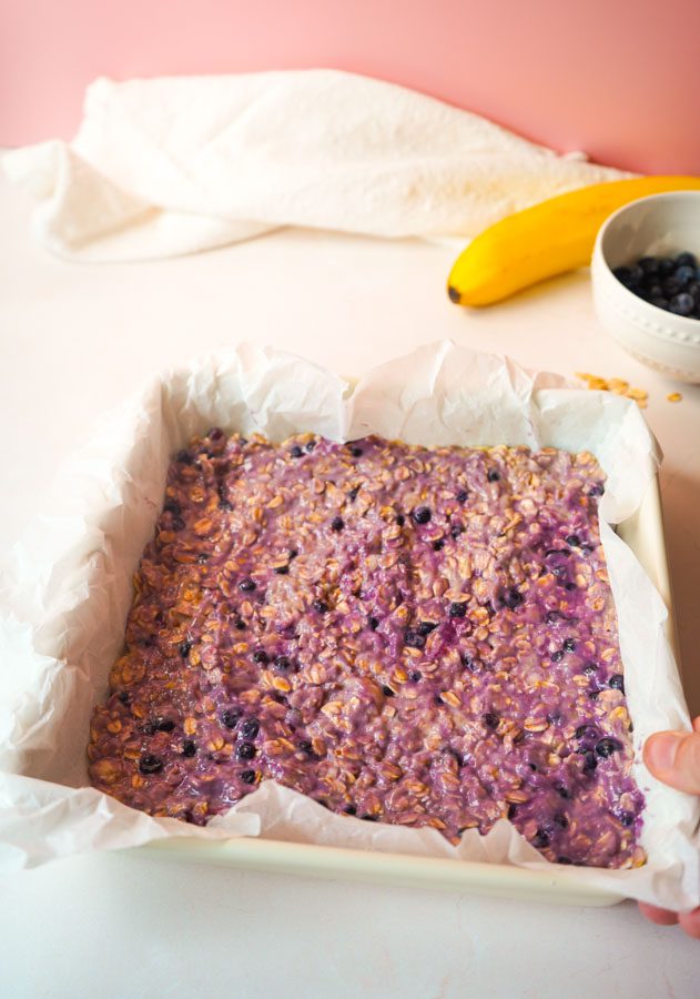Purple blueberry banana oat batter spread evenly in a white parchment lined square baking pan ready to go in the oven with banana and blueberries in the background