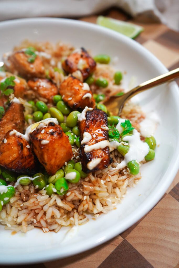 Chipotle honey airfryer salmon bites over brown rice with edamame, lime crema drizzle and sesame seeds in a white bowl