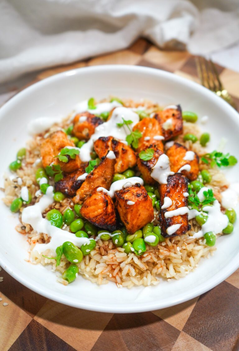 Chipotle honey airfryer salmon bites over brown rice with edamame, lime crema drizzle and sesame seeds in a white bowl