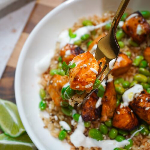 Chipotle honey airfryer salmon bites over brown rice with edamame, lime crema drizzle and sesame seeds in a white bowl