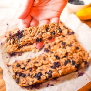 Soft-baked wild blueberry banana oat finger bar broken in half showing a tender, blueberry-studded interior over a parchment paper.
