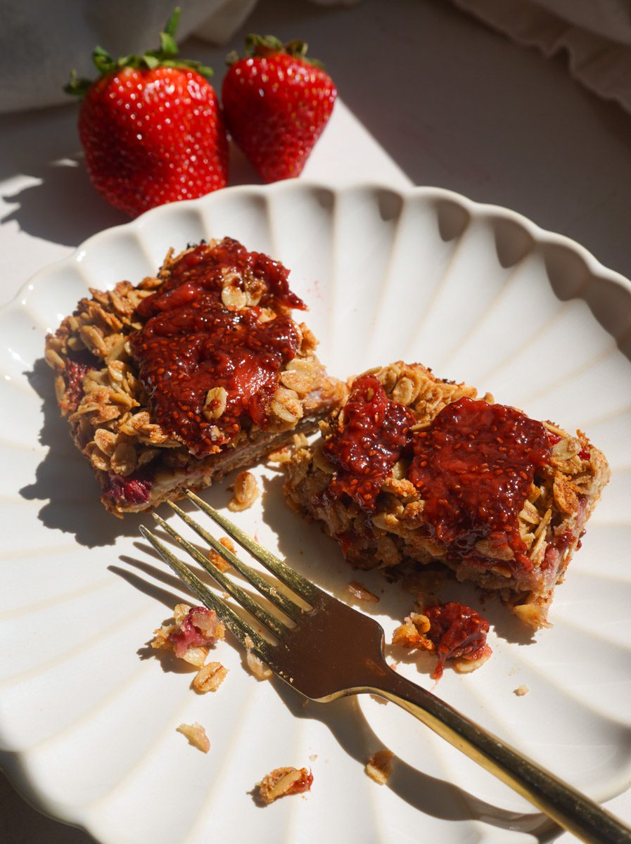 Healthy strawberry chia jam oat squares on a white scalloped plate with yogurt drizzle and gold fork on a pink backdrop