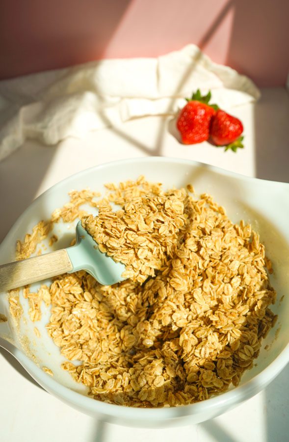 Mixing rolled oats, almond flour, coconut oil and honey for strawberry chia jam oat squares in a white bowl with a light blue spatula and fresh strawberries in the background