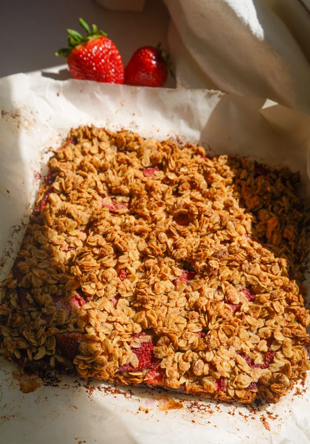 Freshly baked strawberry oat squares on white parchment paper showing golden oat crumble top just out of the oven before the chia jam is added