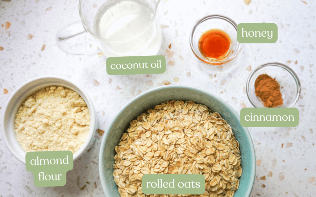 Flat lay ingredients for strawberry chia jam oat squares including rolled oats, almond flour, coconut oil, raw honey and cinnamon in bowls on a white terrazzo surface with sage green ingredient labels