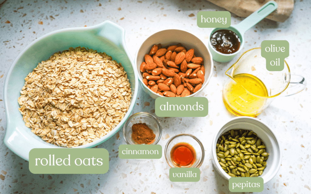 Flat lay of honey roasted almond granola ingredients including rolled oats in a teal bowl, whole almonds, pepitas, olive oil, raw honey, cinnamon and vanilla on a white terrazzo surface