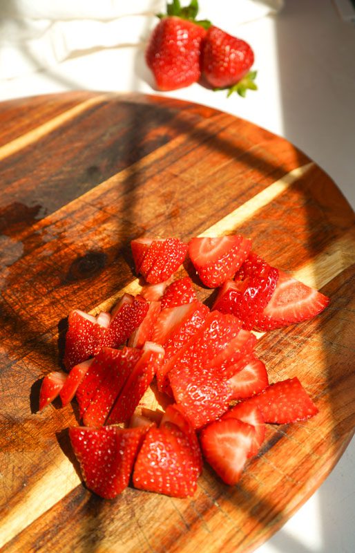 Freshly sliced strawberries on a wooden cutting board with two whole strawberries in the foreground and a white linen napkin
