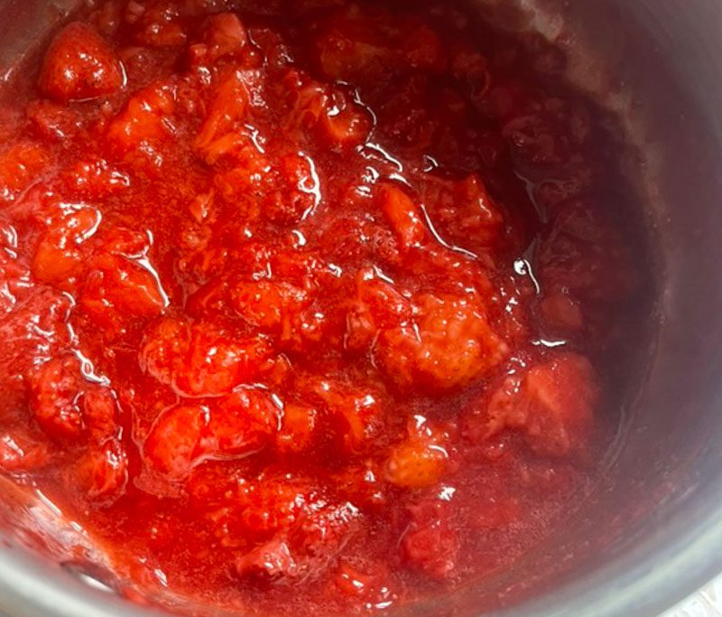 Overhead view of strawberries breaking down and bubbling into a deep red chunky jam in a silver saucepan