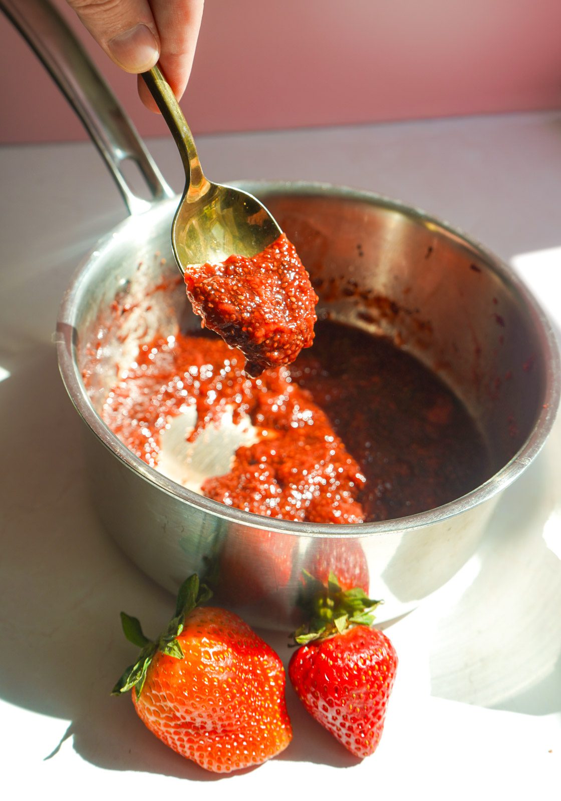 Hand holding a gold spoon lifting thick homemade strawberry chia jam from a silver saucepan with fresh strawberries in the foreground on a pink backdrop