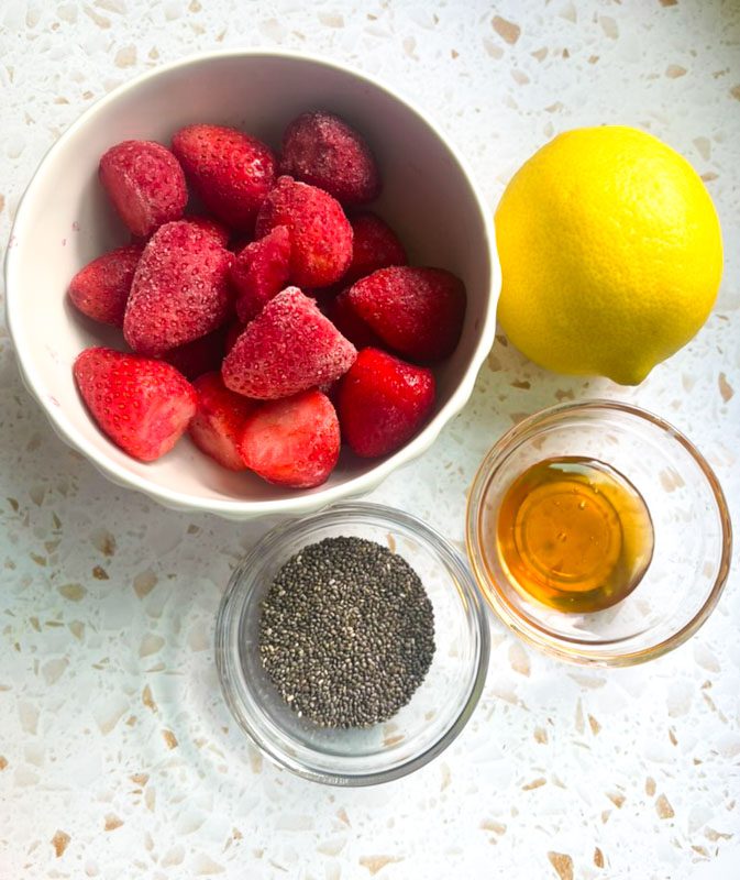 Flat lay of homemade strawberry chia jam ingredients including frozen strawberries in a white bowl, whole lemon, raw honey in a glass bowl and chia seeds on a white terrazzo surface