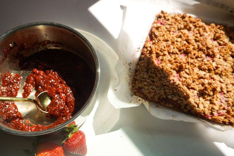 Silver saucepan of homemade strawberry chia jam with gold spoon next to freshly baked strawberry oat squares on parchment paper with fresh strawberries alongside