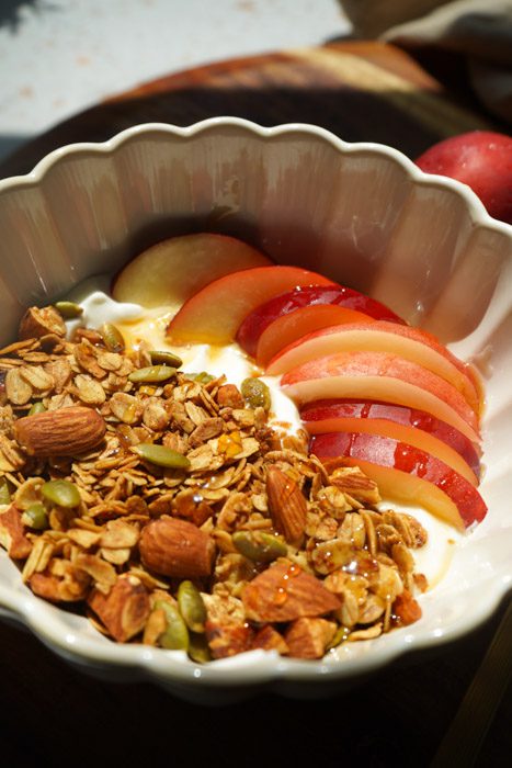 Close up overhead view of honey nectarine yogurt bowl with fresh nectarine slices golden granola honey drizzle and gold spoon in white scalloped bowl on white terrazzo surface