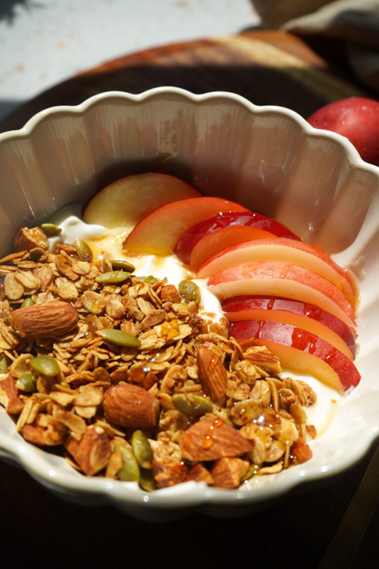 Close up overhead view of honey nectarine yogurt bowl with fresh nectarine slices golden granola honey drizzle and gold spoon in white scalloped bowl on white terrazzo surface