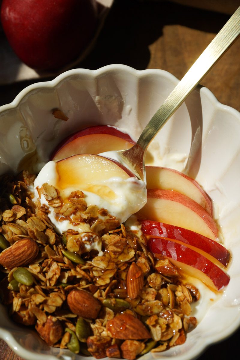 Close up of honey nectarine yogurt bowl in white scalloped bowl showing gold spoon lifting creamy Greek yogurt with honey drizzle fresh nectarine slices and honey roasted almond granola with whole almonds and pepitas