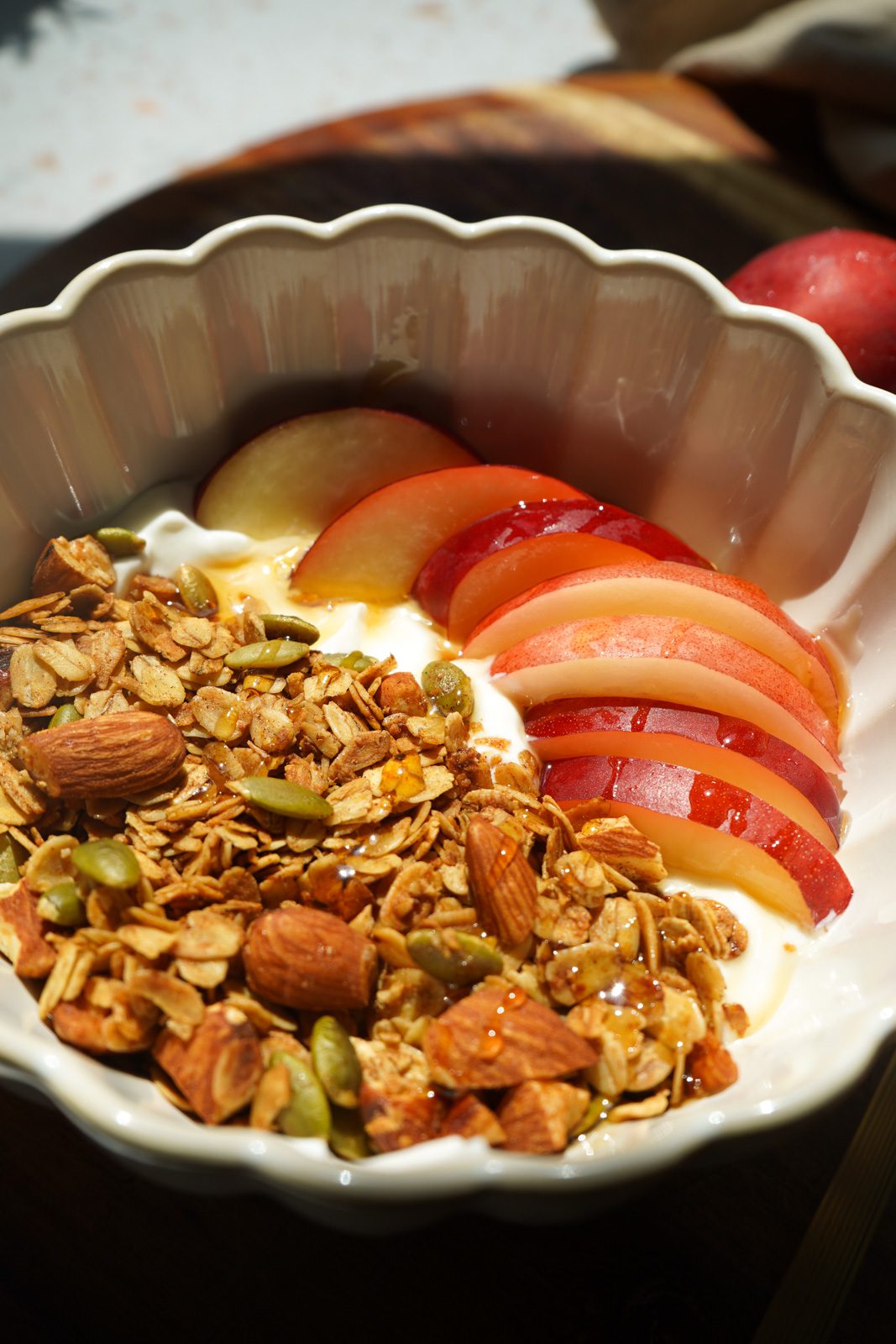 Close up overhead view of honey nectarine yogurt bowl with fresh nectarine slices golden granola honey drizzle and gold spoon in white scalloped bowl on white terrazzo surface